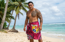 "Tongan man modeling the Fuchsia Kingdom of Tonga Seal Tupenu on a tropical beach. The sarong features the Royal Coat of Arms, intricate tribal patterns, and Tiare flowers."