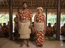 A man and woman standing together in a traditional fale, wearing coordinated outfits made from Tonga Seal Hibiscus floral print fabric. The man wears a button-up shirt and woven ta'ovala, while the woman wears a sleeveless dress with a woven fringe belt and floral lei po’o.