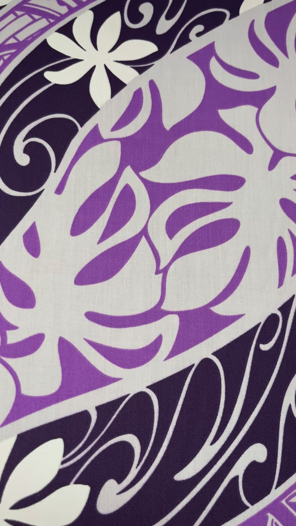 "A close-up video showcasing the 'Tiare Monstera' fabric, featuring a vibrant purple and dark navy wave-print background. The design is layered with white Tiare (gardenia) flowers and purple Monstera leaves, accented by intricate white traditional Polynesian tattoo-style geometric patterns. The video pans across the textile to show the repeating motif and the slight texture of the poly-cotton blend."