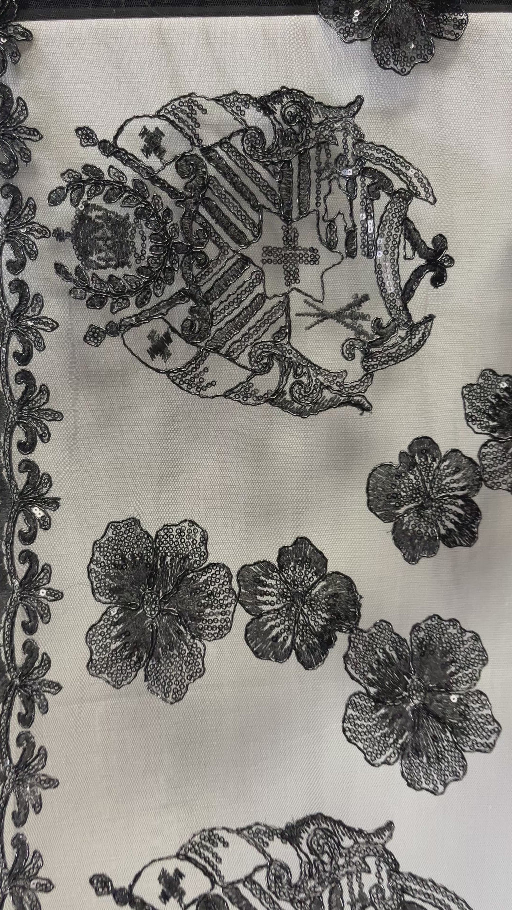 Detailed view of "Hibiscus Tonga Seal Lace" fabric featuring intricate black embroidery of the Tongan Coat of Arms alongside several hibiscus blossoms with sequin accents on a sheer white base.
